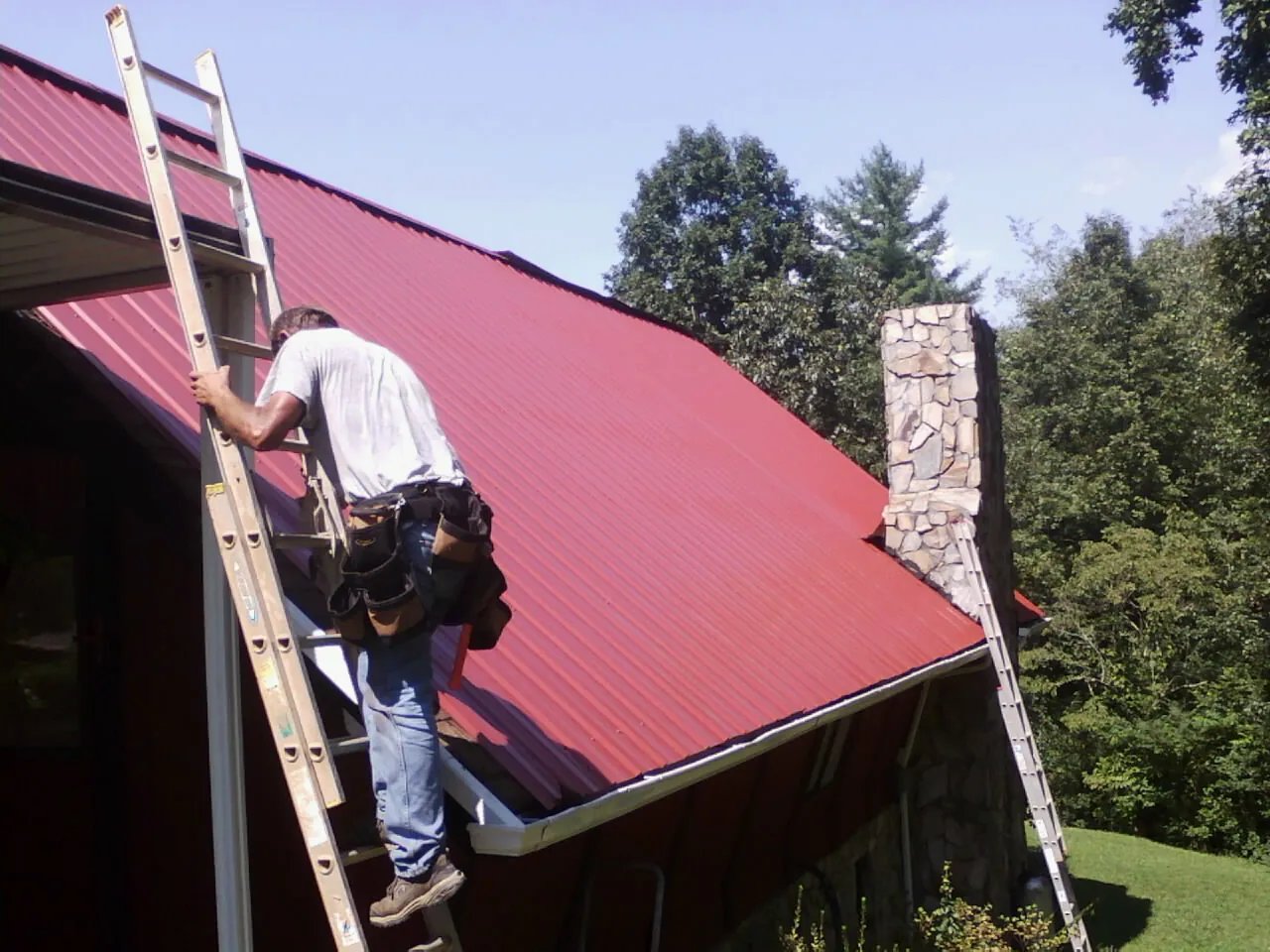 Roofer on a ladder beside a red metal roof and stone chimney — Soffit & Fascia Repair by Roofing Brothers in Scott Depot